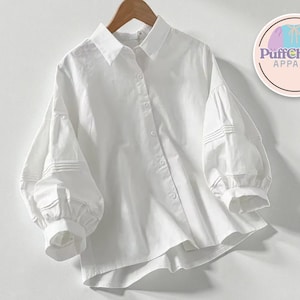 May include: A white button-down shirt with long, voluminous sleeves. The shirt has a classic collar and button-up front. The sleeves have pleated details near the cuffs. The PuffCharm Apparel logo is visible in the upper right corner.