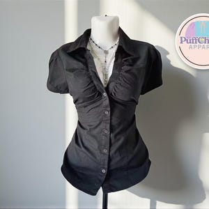 Puede incluir: Camisa negra abotonada con mangas cortas abullonadas. La camisa tiene cuello y una silueta entallada. Se ve un collar en el escote. La camisa se exhibe en un maniquí. El logotipo de PuffCharm Apparel está en el fondo.
