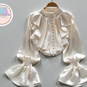 May include: Ivory-colored blouse with a high collar, ruffled front, and long, flared sleeves. The blouse has a row of small buttons down the front and elastic cuffs. The PuffCharm Apparel logo is in the upper left corner.