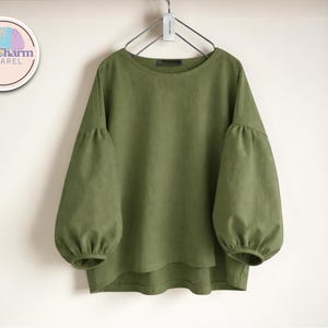 May include: Olive green long-sleeved top with a round neckline and voluminous sleeves. The top is hanging on a metal hanger. The PuffCharm Apparel logo is visible in the upper left corner.