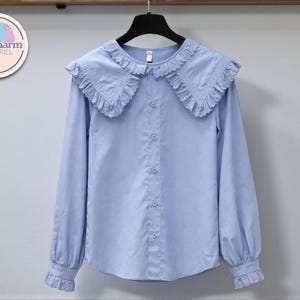 May include: A light blue long-sleeve blouse with a ruffled collar and cuffs. The blouse has a button-down front and is hanging on a black hanger. The PuffCharm Apparel logo is in the upper left corner.