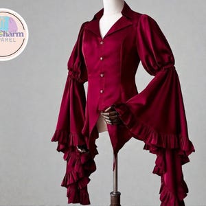 Op de afbeelding: Bordeaux fluwelen blouse met een V-hals, knoopsluiting en dramatische klokmouwen. De mouwen zijn geaccentueerd met ruches bij de elleboog en pols. De blouse wordt op een mannequin getoond.