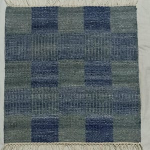 May include: A square, handwoven rug with a checkered pattern in shades of blue and gray. The rug has a textured surface and a fringe of white tassels along the top and bottom edges. The rug is suitable for indoor use.