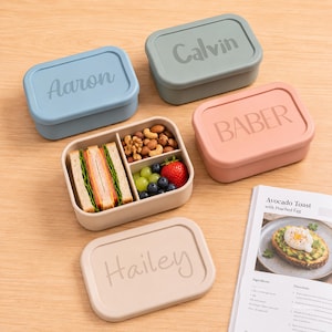 Op de afbeelding: Een verzameling gepersonaliseerde lunchboxen in verschillende kleuren, waaronder blauw, groen en perzik. Eén doos is open en toont sandwiches, noten en vers fruit. Elke deksel is gegraveerd met een naam: Aaron, Calvin, Baber en Hailey.