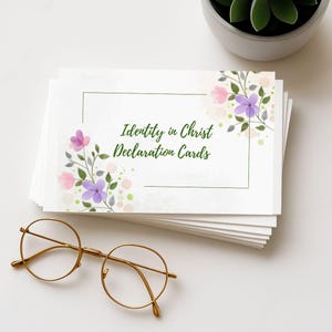 May include: A stack of white cards with floral accents and the text "Identity in Christ Declaration Cards" in green. The cards are decorated with pink and purple flowers. Gold-rimmed glasses rest in front of the cards.