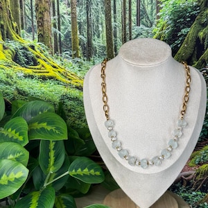 May include: A necklace featuring a gold-toned chain with a series of pale green, frosted, cube-shaped beads interspersed with small white pearls and tiny gold-toned spacer beads, displayed on a neutral-coloured jewellery bust.