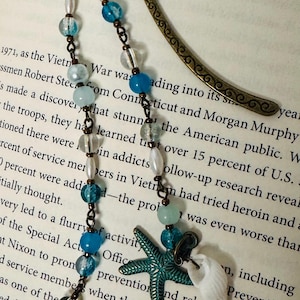 Puede incluir: Un marcapáginas con cuentas con una parte superior de color bronce y una cadena decorativa. La cadena presenta cuentas azules, transparentes y blancas, junto con una estrella de mar turquesa y un dije de concha blanca. El marcapáginas está sobre un libro.