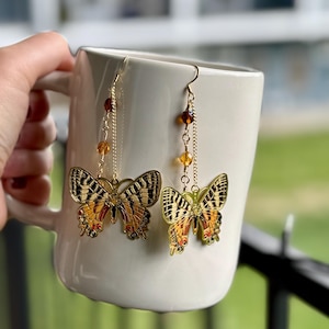 以下が含まれることがあります： 金色の装飾が施された蝶のイヤリングのペア。 蝶の羽は、黒、オレンジ、黄色の詳細なパターンが特徴です。 イヤリングは、琥珀色のビーズが付いた金色のフックとチェーンから吊り下げられています。