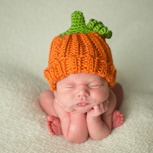 May include: A baby wearing an orange and green knitted pumpkin hat. The baby is lying on a white blanket and has their eyes closed.