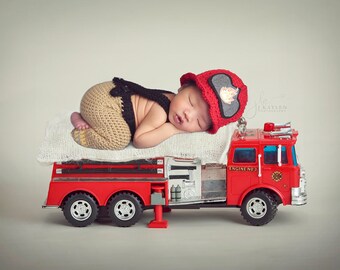 May include: A red toy fire truck with a white blanket on top. A baby wearing a red firefighter hat and brown pants is sleeping on the blanket.