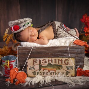 May include: A newborn baby wearing a brown and white crocheted fishing outfit sleeps in a wooden crate with a fishing sign that reads "Fishing Expeditions" and a fishing pole. The crate is surrounded by fall leaves and a small metal bucket.