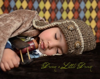 May include: A young child wearing a brown crocheted hat with white trim and brown buttons sleeps soundly while holding a wooden toy airplane. The child is lying on a brown and yellow patterned blanket.