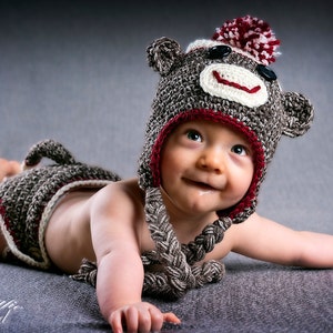 May include: A baby wearing a brown and red crocheted monkey hat with a white face and black eyes. The baby is lying on a gray surface and looking up.