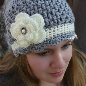 May include: A gray crocheted beanie with a white crocheted flower and a white crocheted band around the brim.