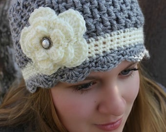 May include: A gray crocheted beanie with a white crocheted flower and a white crocheted band around the brim.