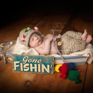 May include: A baby wearing a crocheted fishing hat and overalls is lying in a blue and white boat-shaped basket. The basket is decorated with a sign that says "Gone Fishin'" and there are colorful crocheted fish hanging from the side.