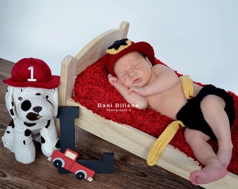 May include: A newborn baby wearing a red and yellow firefighter hat and matching outfit is sleeping on a red and white blanket in a wooden crib. A plush dalmatian dog with a red fire hat is on the left side of the image. A black letter L with a red toy truck is on the right side of the image. The text "Dani Dillena Photography ©" is visible in the image.