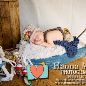 May include: A baby boy wearing a brown and white knit hat and a brown and white knit outfit is sleeping in a blue and white wooden boat. The boat is decorated with a fishing net and a red and white fishing bobber. The baby is surrounded by a white fluffy blanket.