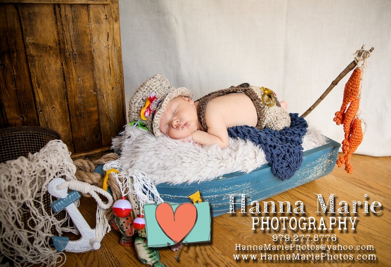 May include: A baby boy wearing a brown and white knit hat and a brown and white knit outfit is sleeping in a blue and white wooden boat. The boat is decorated with a fishing net and a red and white fishing bobber. The baby is surrounded by a white fluffy blanket.
