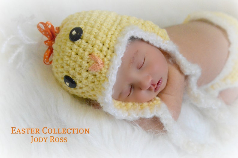 Op de afbeelding: Een gele gehaakte muts met zwarte knoopogen, een kleine oranje geborduurde snavel en een oranje strik met stippen bovenop. De muts heeft een witte gehaakte rand en lange oorflappen. Tekst: EASTER COLLECTION JODY ROSS.