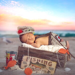 May include: A sleeping baby wearing a crocheted fishing hat and overalls is lying in a wooden crate with a sign that says "Fishing" and a fish illustration. The crate is on a sandy beach with seashells and a bucket.