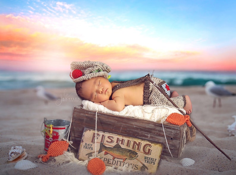 May include: A sleeping baby wearing a crocheted fishing hat and overalls is lying in a wooden crate with a sign that says "Fishing" and a fish illustration. The crate is on a sandy beach with seashells and a bucket.