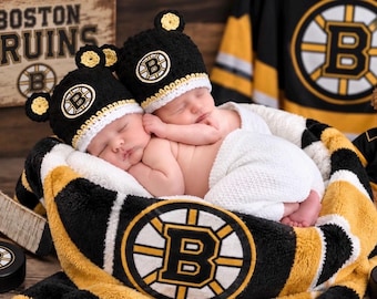 May include: Two infants wearing black crocheted beanies with yellow bear ears and the Boston Bruins logo, wrapped in a white blanket. The set includes a black and yellow Boston Bruins patterned blanket, a hockey stick, and a teddy bear.