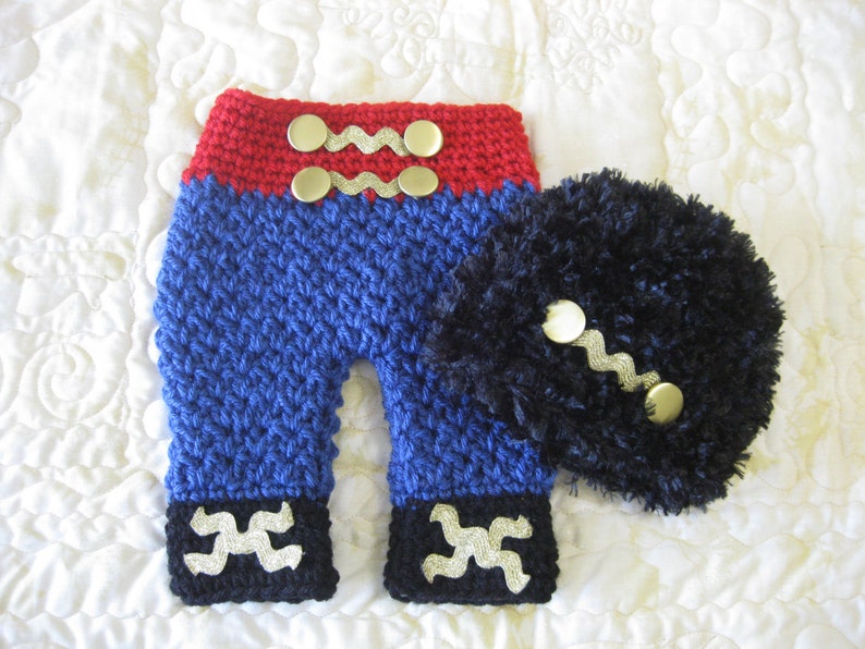 Puede incluir: Conjunto de beb&eacute; de ganchillo en rojo, azul y negro con botones dorados. El conjunto incluye pantalones y un gorro.
