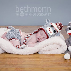May include: A baby wearing a gray and red crocheted monkey hat and matching outfit sleeps on a white blanket next to a gray sock monkey toy.