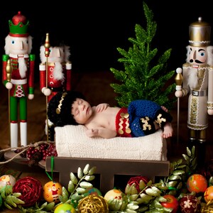 Puede incluir: Un beb&eacute; reci&eacute;n nacido vestido con un atuendo de crochet azul y rojo est&aacute; durmiendo en un trineo de madera rodeado de adornos navide&ntilde;os y figuras de cascanueces.