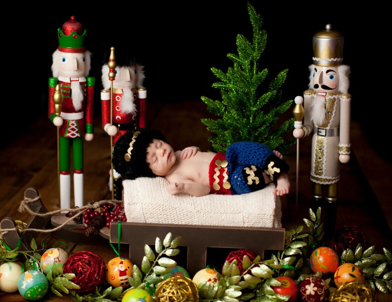Puede incluir: Un beb&eacute; reci&eacute;n nacido vestido con un atuendo de crochet azul y rojo est&aacute; durmiendo en un trineo de madera rodeado de adornos navide&ntilde;os y figuras de cascanueces.