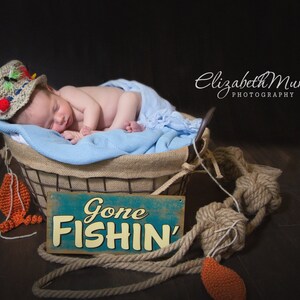 May include: A newborn baby wearing a crocheted fishing hat sleeps in a wicker basket lined with a blue blanket. The basket is surrounded by a rope and a crocheted orange fish. A sign reads "Gone Fishin'"