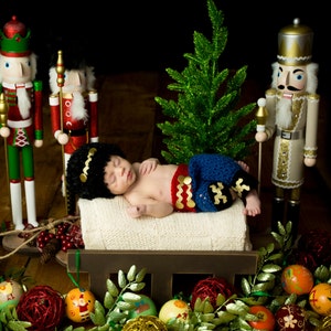 Puede incluir: Un beb&eacute; vestido de cascanueces est&aacute; durmiendo en un trineo de madera rodeado de adornos navide&ntilde;os y figuras de cascanueces. El beb&eacute; lleva un atuendo de crochet azul y rojo con un sombrero negro. El trineo est&aacute; decorado con vegetaci&oacute;n y bayas rojas.