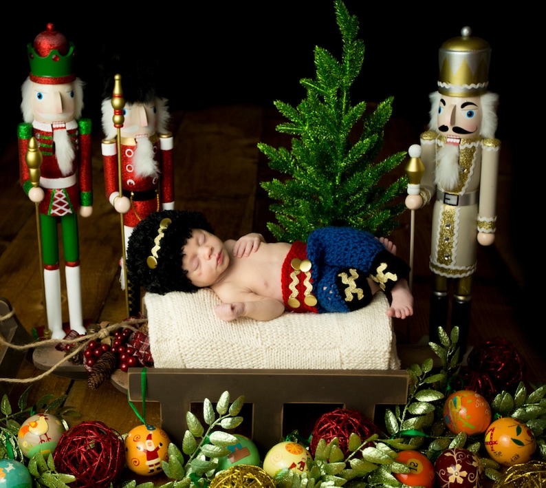Puede incluir: Un beb&eacute; vestido de cascanueces est&aacute; durmiendo en un trineo de madera rodeado de adornos navide&ntilde;os y figuras de cascanueces. El beb&eacute; lleva un atuendo de crochet azul y rojo con un sombrero negro. El trineo est&aacute; decorado con vegetaci&oacute;n y bayas rojas.
