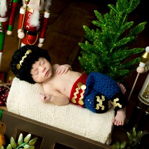 Puede incluir: Un beb&eacute; reci&eacute;n nacido lleva un atuendo de crochet rojo y azul con detalles dorados, durmiendo en una manta blanca. El beb&eacute; lleva un gorro de punto negro con una banda dorada. El beb&eacute; est&aacute; rodeado de decoraciones navide&ntilde;as, incluyendo un cascanueces y un peque&ntilde;o &aacute;rbol de Navidad artificial.
