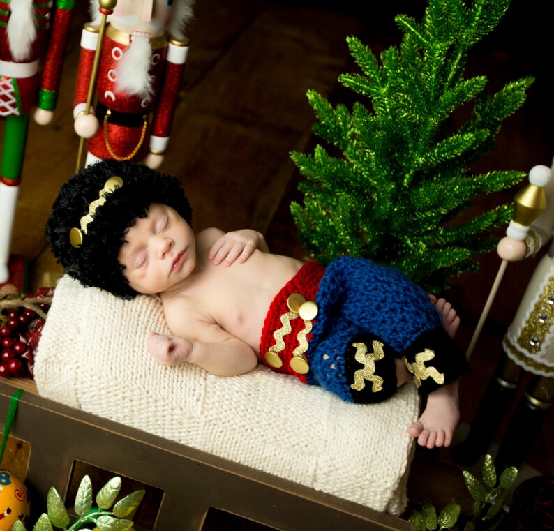 Puede incluir: Un beb&eacute; reci&eacute;n nacido lleva un atuendo de crochet rojo y azul con detalles dorados, durmiendo en una manta blanca. El beb&eacute; lleva un gorro de punto negro con una banda dorada. El beb&eacute; est&aacute; rodeado de decoraciones navide&ntilde;as, incluyendo un cascanueces y un peque&ntilde;o &aacute;rbol de Navidad artificial.