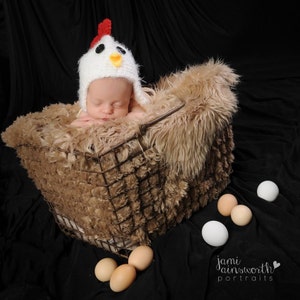 May include: A baby wearing a white crocheted chicken hat is lying in a brown faux fur-lined wire basket.  There are several white eggs scattered around the basket.