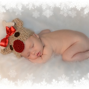 May include: A newborn baby wearing a brown and red reindeer hat with a red bow sleeps on a white fluffy blanket with a white snowflake border.