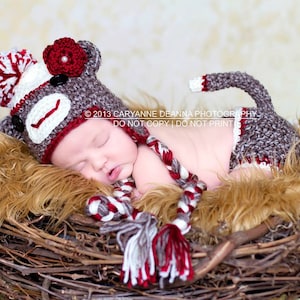 May include: A newborn baby wearing a crocheted monkey hat and matching diaper cover. The hat has a red flower and white pom-pom. The baby is sleeping in a nest made of twigs.