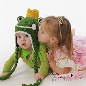 May include: A young child wearing a green frog hat with a gold crown and a white brim is being kissed on the cheek by another young child wearing a pink dress and a sparkly tiara.