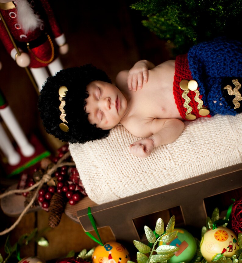 Puede incluir: Un beb&eacute; durmiendo con un atuendo de punto rojo y azul con un gorro de piel negro, acostado sobre una manta blanca en una cuna de madera. La cuna est&aacute; decorada con follaje verde y huevos de Pascua coloridos.