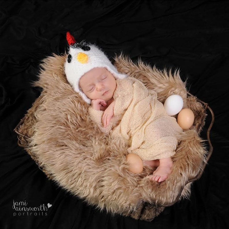 May include: A baby wearing a white crocheted chicken hat sleeps in a brown faux fur nest with three white and brown eggs.