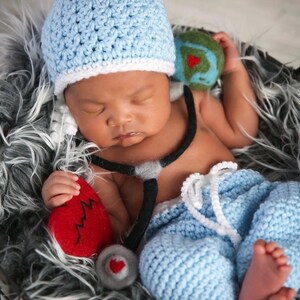 May include: A light blue crocheted beanie with a white brim, light blue crocheted pants with a white drawstring, a black and gray felt stethoscope prop, a red felt heart with a black zigzag line, and a small green and blue felt globe.