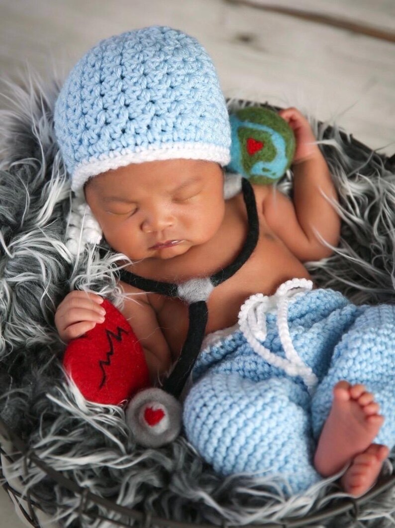 May include: A light blue crocheted beanie with a white brim, light blue crocheted pants with a white drawstring, a black and gray felt stethoscope prop, a red felt heart with a black zigzag line, and a small green and blue felt globe.