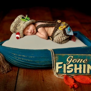 May include: A newborn baby wearing a brown and green crocheted fishing outfit is sleeping in a blue wooden boat. The boat is on a wooden surface with a sign that says "Gone Fishin'" in yellow letters.