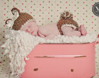 May include: A newborn baby wearing a brown and white crocheted hat with ears and a matching diaper cover with a pom-pom, lying in a pink metal tub. The baby is sleeping on a white, fluffy blanket. The background is a polka dot pattern.