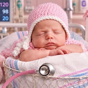 May include: A sleeping infant wearing a pink crocheted hat with white trim, resting in a basket lined with a pink and blue striped blanket. A pink teddy bear with a Tiny Fighter tag and a pink stethoscope are placed near the infant.