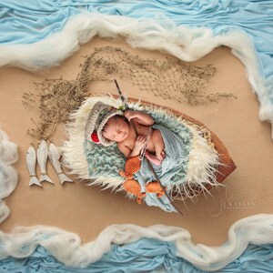May include: A newborn baby wearing a crocheted hat and a blue blanket is lying in a wooden boat. The boat is surrounded by a blue and white fabric that resembles waves. There are wooden fish and a fishing net in the background.