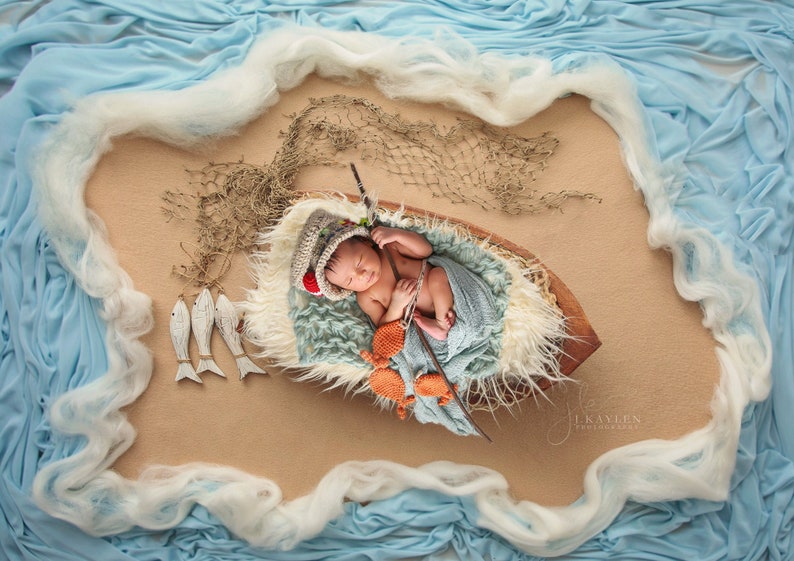 May include: A newborn baby wearing a crocheted hat and a blue blanket is lying in a wooden boat. The boat is surrounded by a blue and white fabric that resembles waves. There are wooden fish and a fishing net in the background.