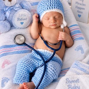 May include: A newborn wearing a light blue crocheted hat and pants, lying on a white blanket with blue and pink stripes. A blue stethoscope rests on the infant. A light blue teddy bear with a Little Patient tag and a sign reading Tiny Miracles Happen Here are visible.
